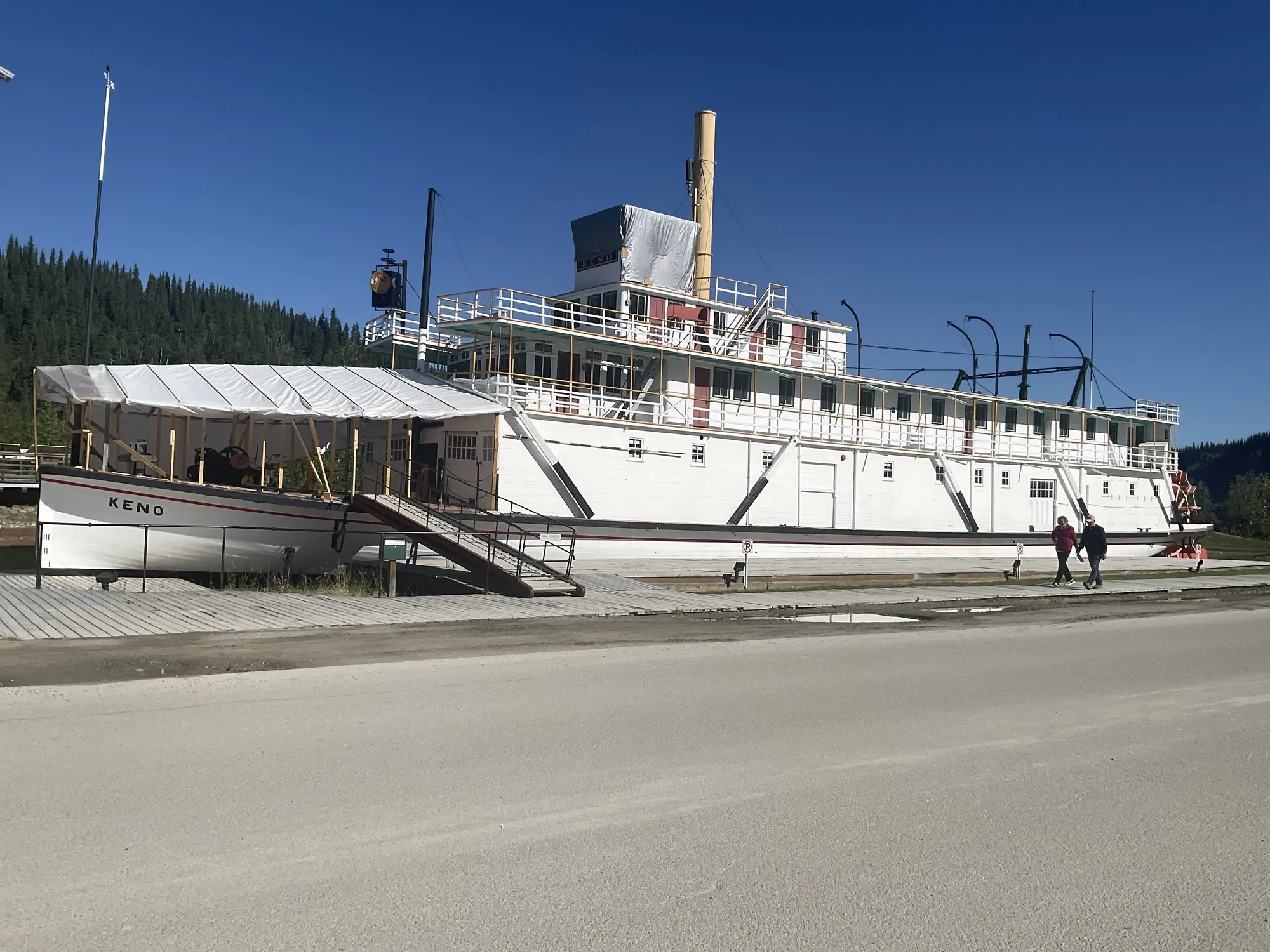 The Keno on blocking beside the street in Dawson City, white hull and decks with a buff stack, the forward main deck under a temporary shelter, forested hill behind