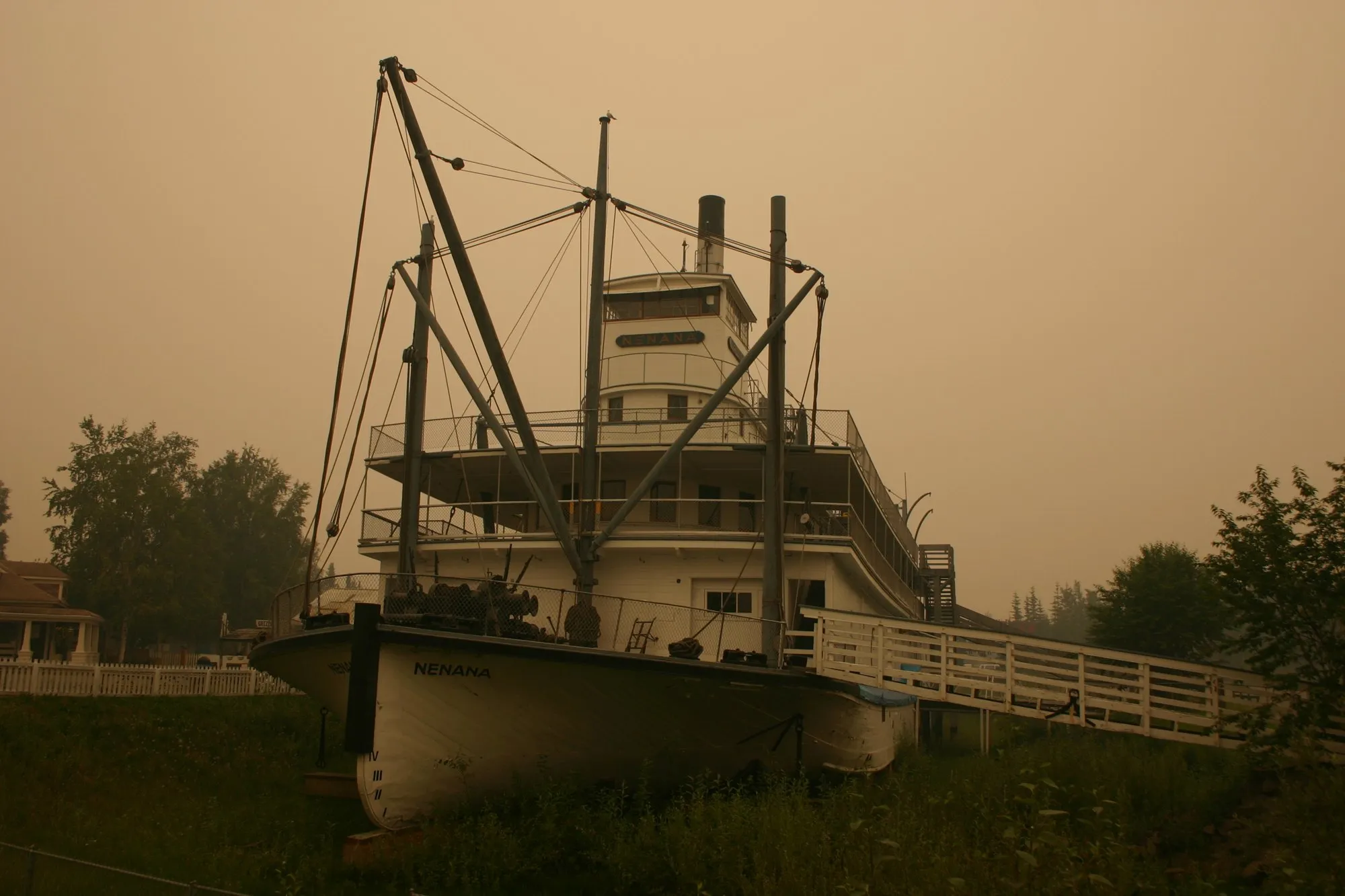The sternwheeler Nenana on display, white hull and three decks under a smoke-hazed amber sky, with spars and rigging at the bow